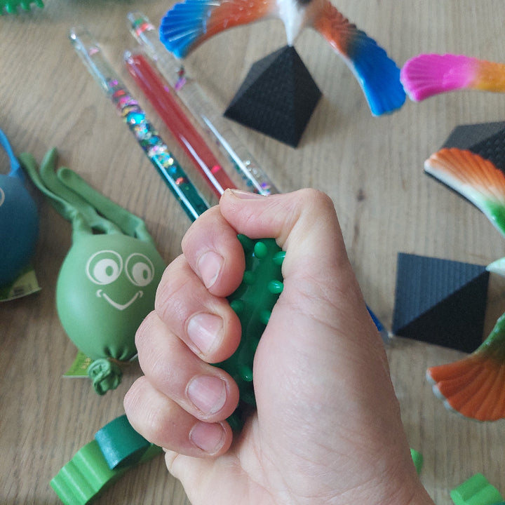 Hand knijpt in een massage-olijf met voelbare stekeltjes voor sensorische afleiding tijdens een medische behandeling, met de overige materialen uit de set op de achtergrond.